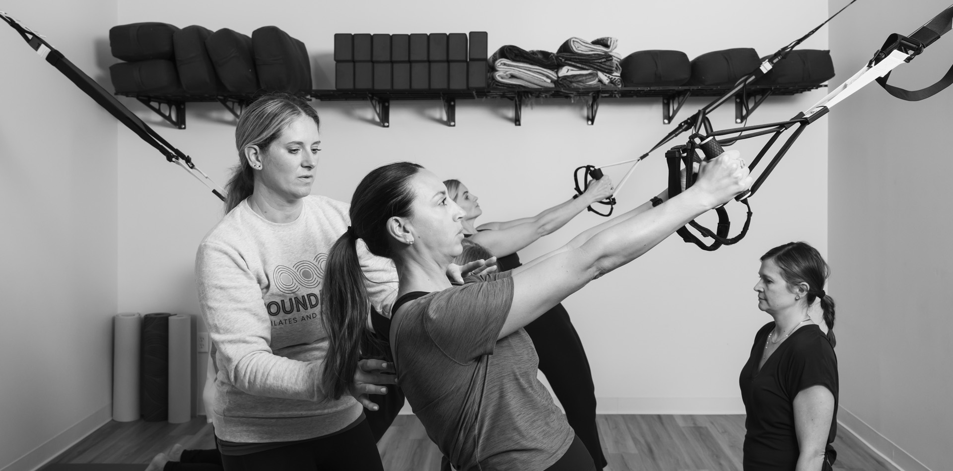 Mat Pilates Instructor supporting a client's posture during a suspension strap exercise in a group class, with shelved Pilates props in the background.