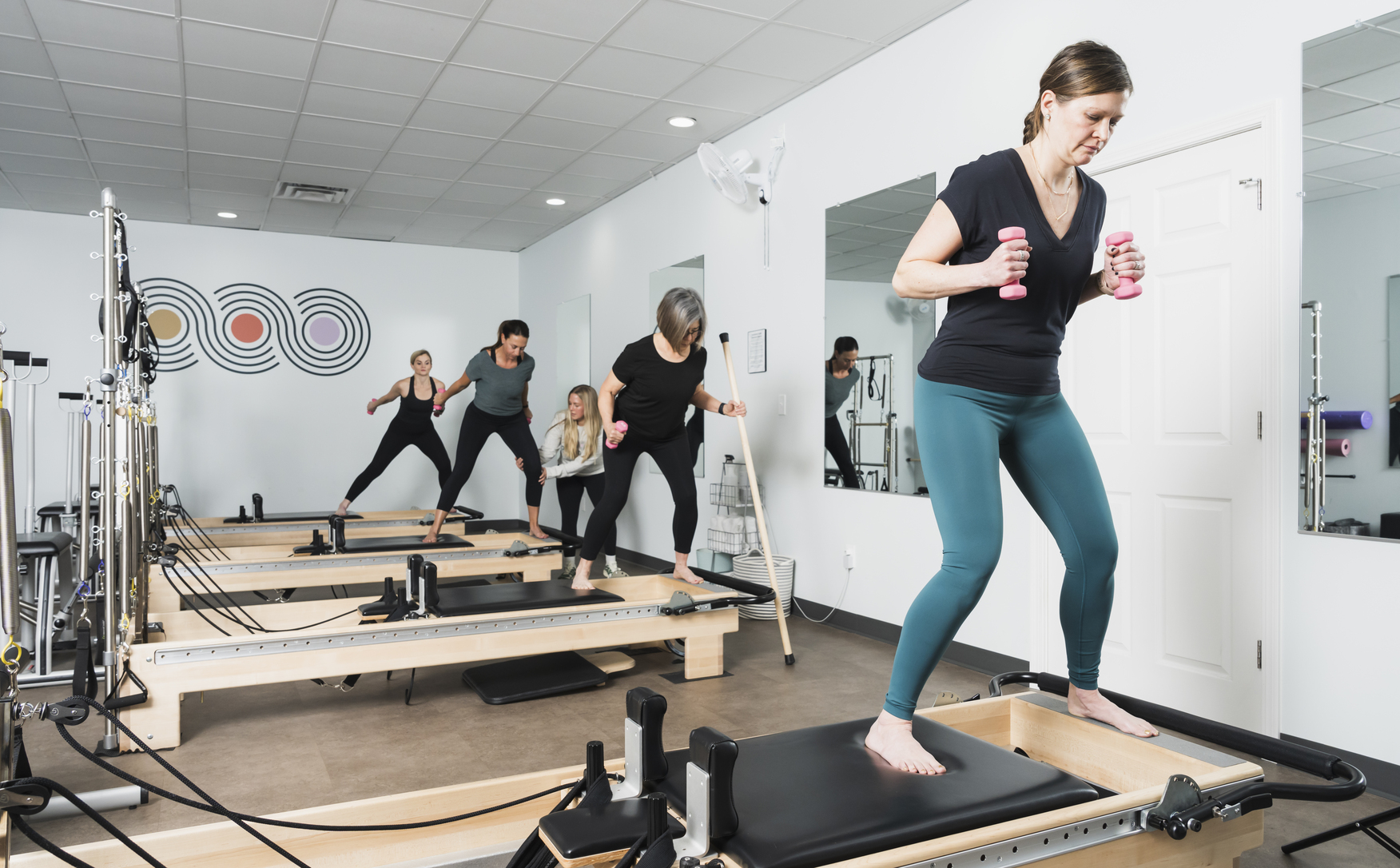 Group reformer Pilates class with participants balancing on reformers and using small hand weights, led by an instructor in a studio with mirrored walls.