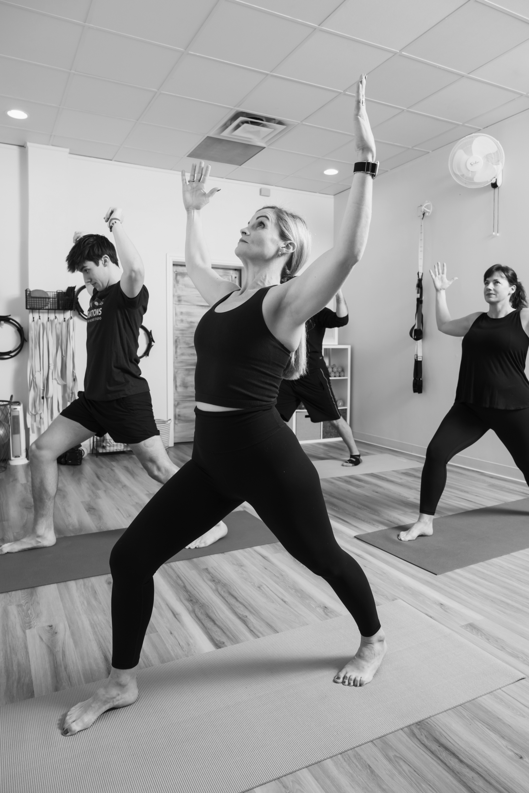 Group of participants in a mat Pilates class holding a wide-legged lunge with arms raised overhead in a studio.