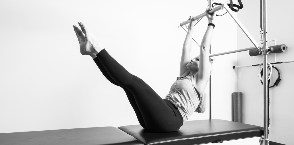 Woman doing a leg-lift exercise on a Pilates Cadillac apparatus, holding the overhead bar with legs extended.