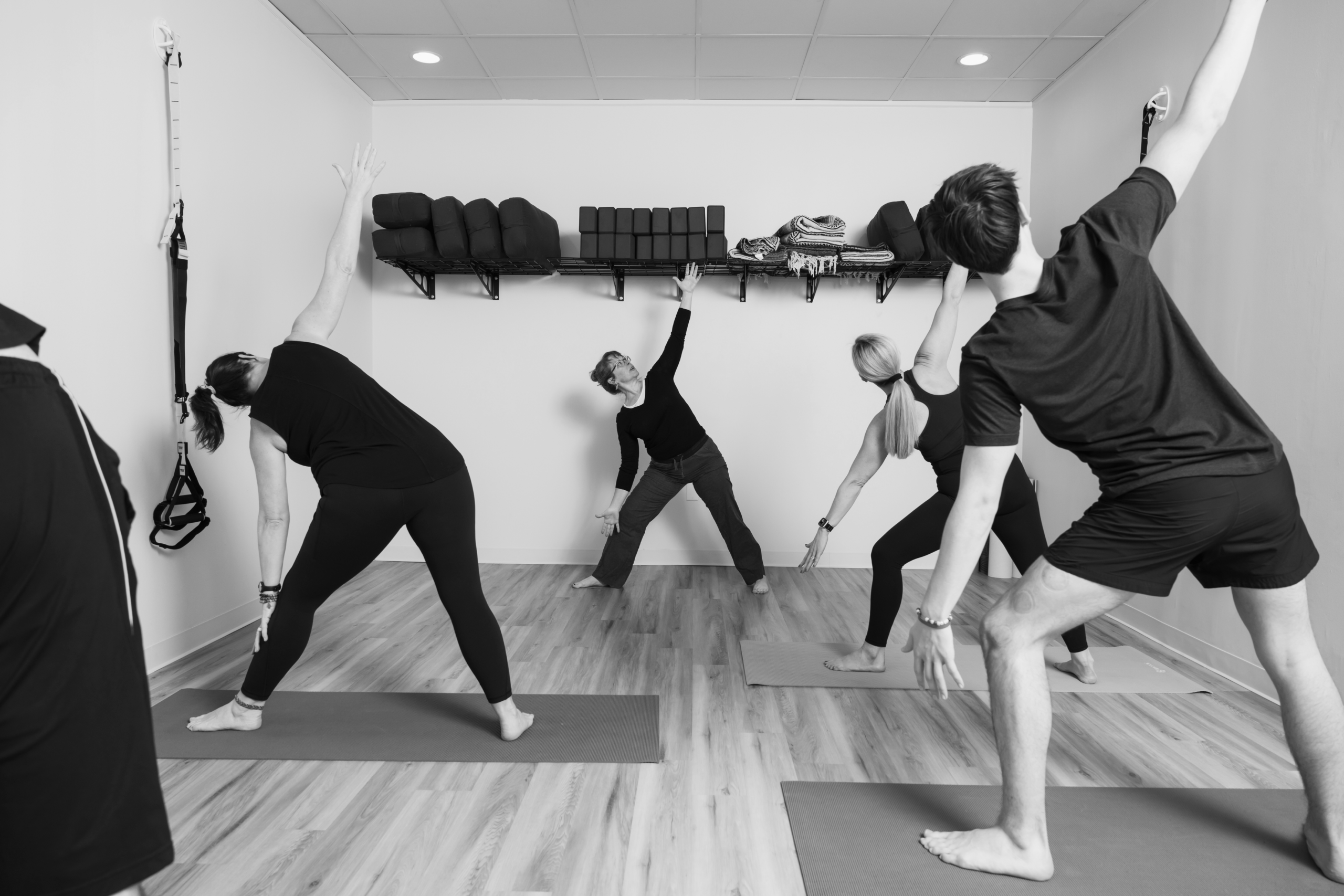 Mat Pilates class holding a triangle-style side stretch with one arm reaching overhead in a studio with shelved equipment.