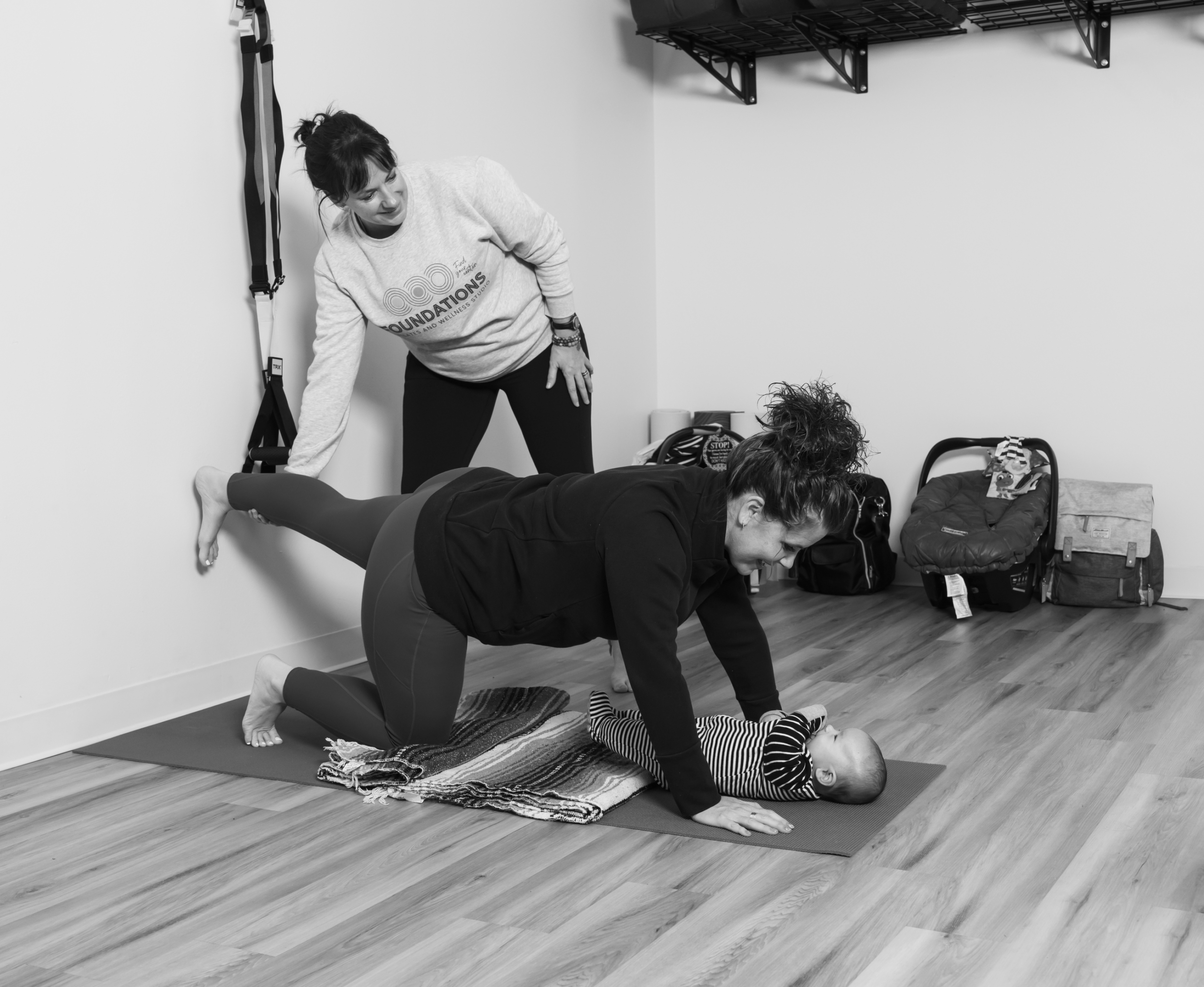 Foundations Studio postpartum pilates instructor supervising a mother performing a quadruped leg lift over her baby on a yoga mat