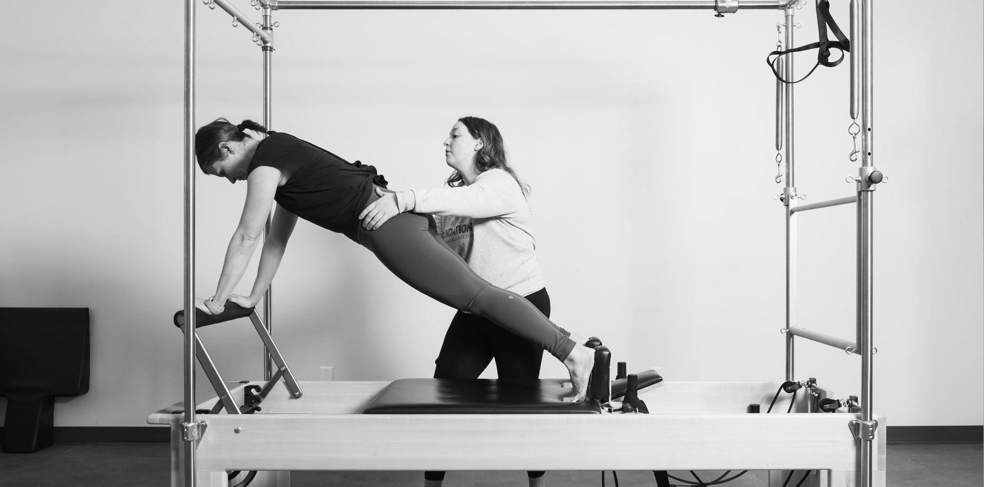 Pilates instructor supporting a client's hips through a plank-style stretch on a Cadillac apparatus.