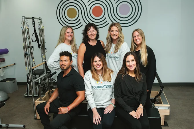 Group portrait of seven smiling Foundations Studio staff members posing together in front of the studio's logo wall.