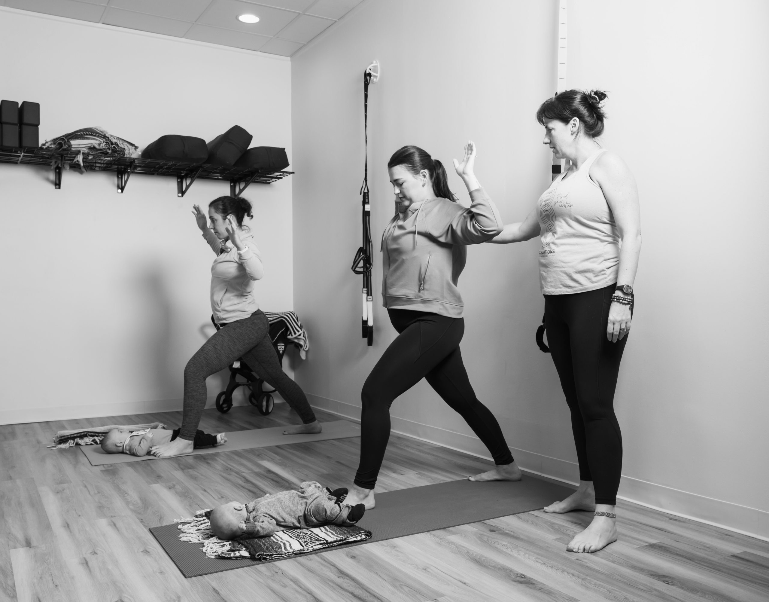 Foundations Studio postpartum pilates instructor providing hands-on guidance to a mother in a warrior pose, with a second mother practicing in the background and babies resting on yoga mats nearby