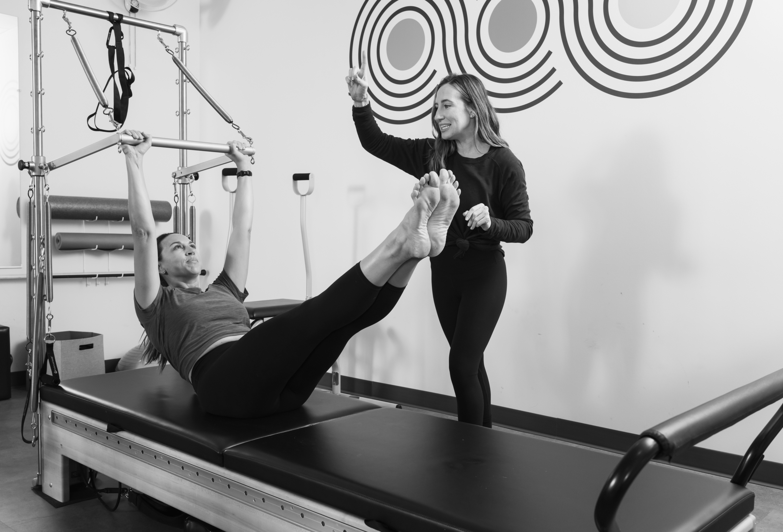 Pilates instructor guiding a client through a reformer exercise, with the client lying back gripping the push-through bar and legs extended.